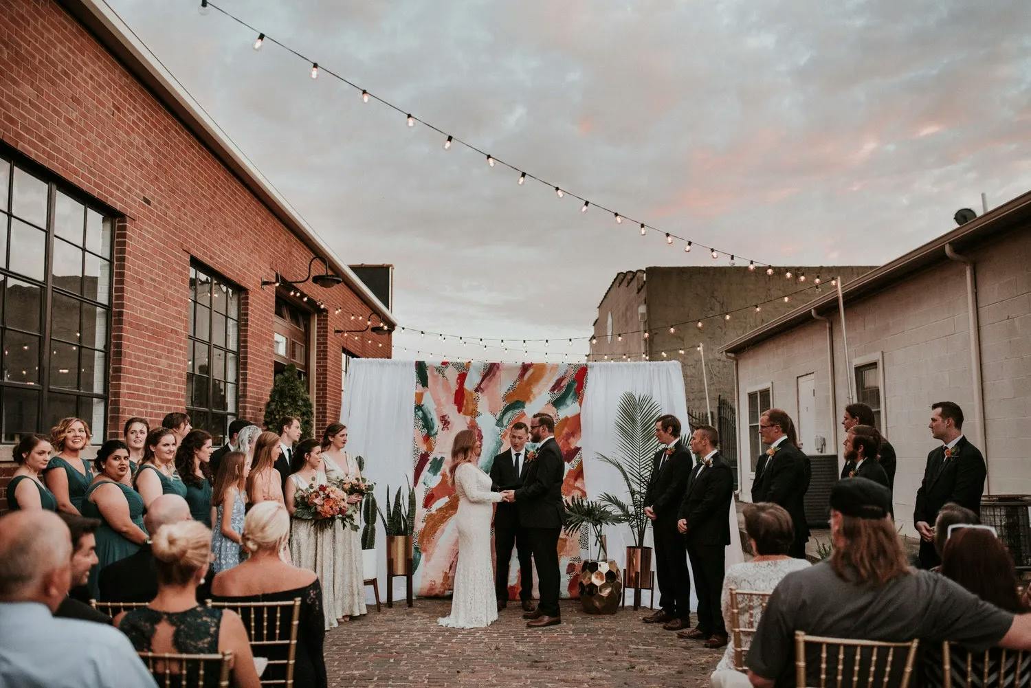 A couple stands facing each other under a colorful abstract backdrop during an outdoor wedding ceremony. They are surrounded by their wedding party, guests in chairs, with string lights overhead. The scene is set between two brick and white buildings at sunset.