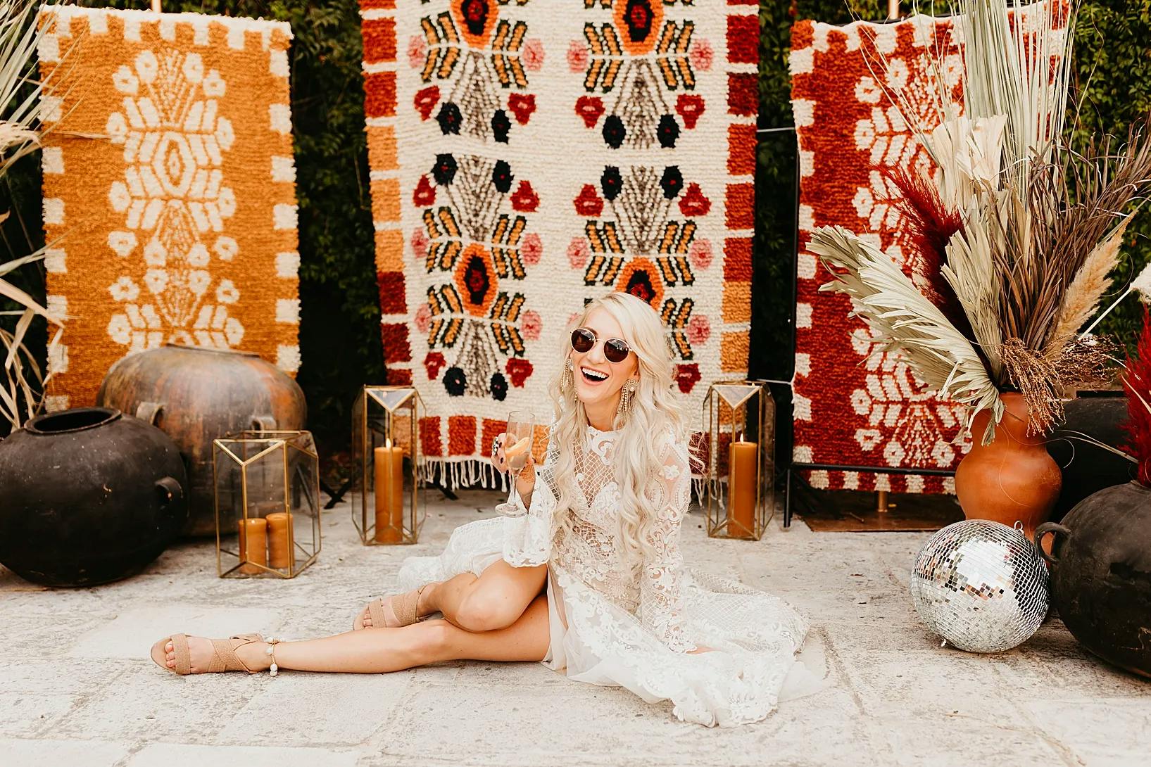 A woman with long blonde hair and sunglasses is sitting on the ground, smiling. She’s wearing a white lace outfit and holding a drink. Behind her are colorful, patterned rugs hanging against a green hedge, with vases, dried plants, and candles placed around her.