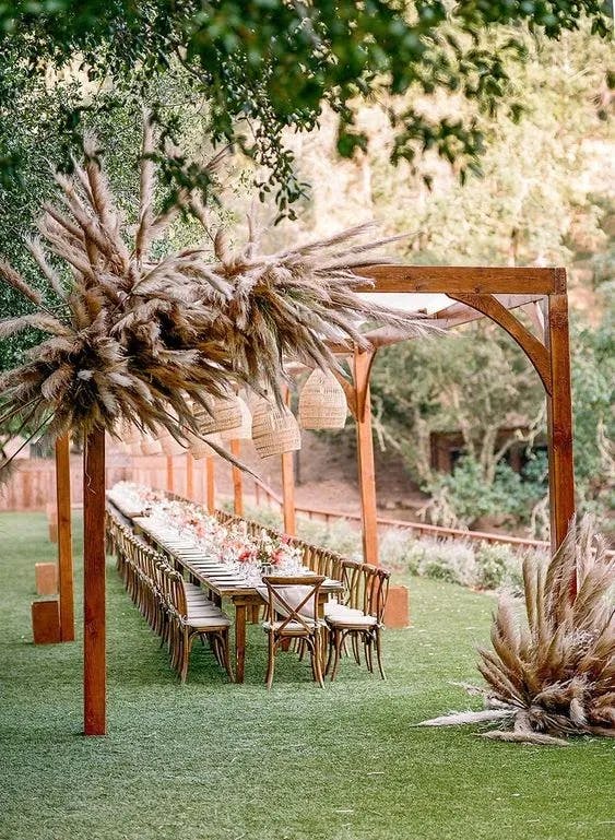 An outdoor dining setting featuring a long wooden table adorned with white flowers and surrounded by wooden chairs. The setup is under a wooden pergola decorated with large pampas grass arrangements and hanging woven lanterns, set on a lush green lawn.