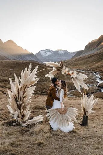 A couple is kissing outdoors in a mountainous landscape at sunset. They are surrounded by pampas grass decorations, with snow-capped peaks and a gentle stream in the background. The bride wears a white dress and holds a circular fan, while the groom dons a brown suit.