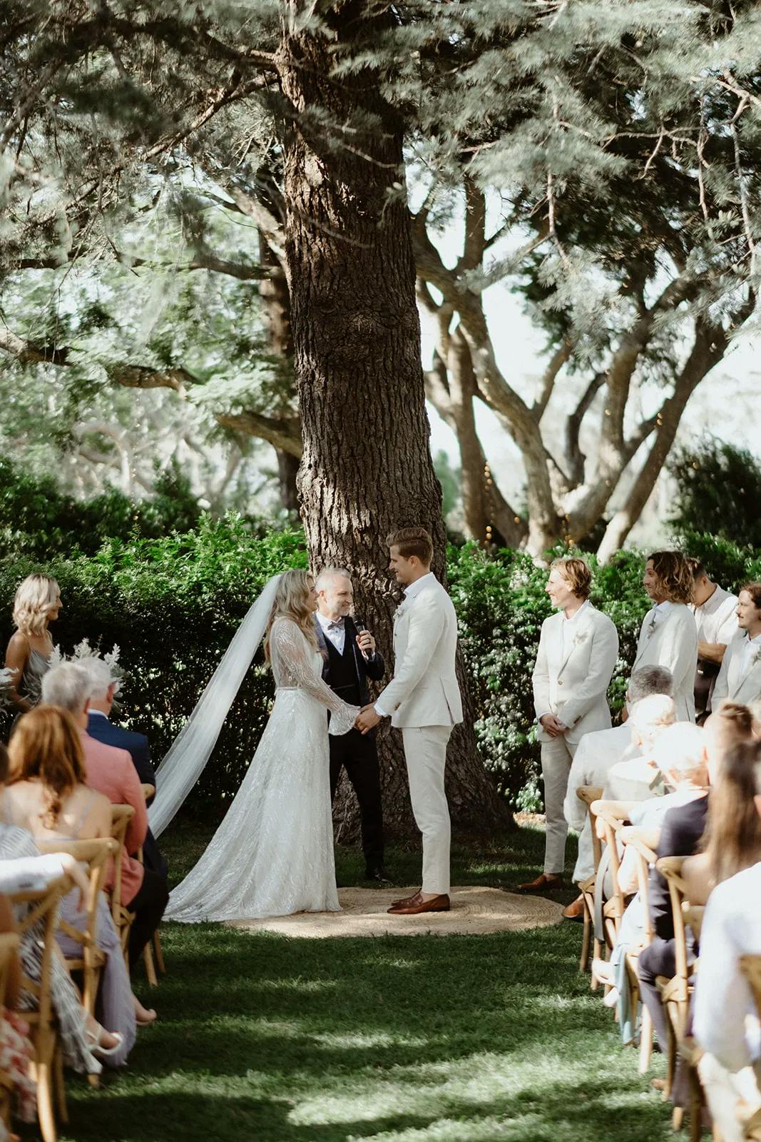 A bride and groom stand before an officiant, holding hands under a large tree during their outdoor wedding ceremony. Bridesmaids and groomsmen dressed in light-colored attire stand on either side, and guests are seated, watching attentively in the background.