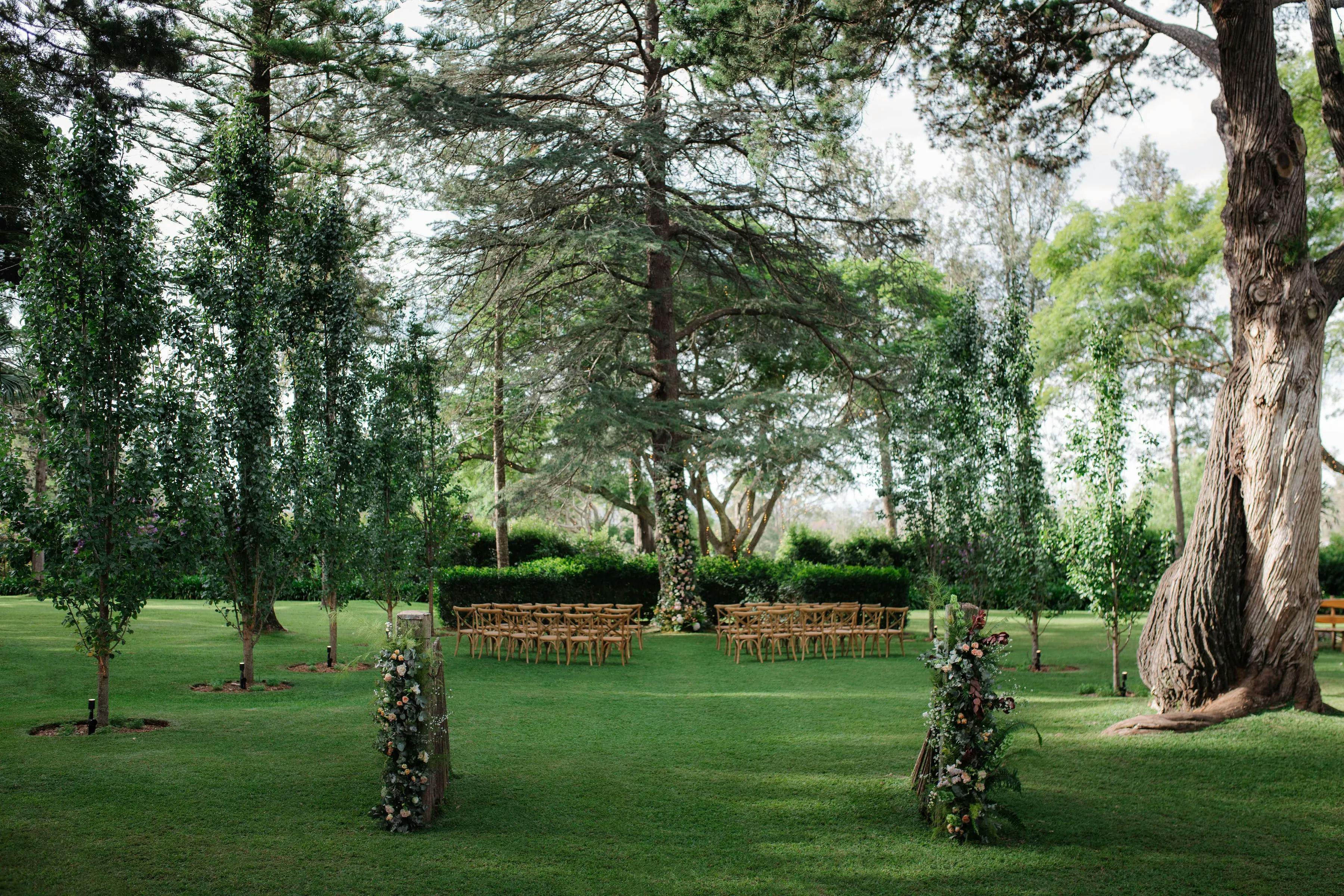 A serene outdoor wedding setup with wooden chairs arranged in rows facing forward under large trees. The grassy aisle is flanked by tall, sparse trees and floral arrangements, leading to an altar area adorned with flowers, set against a backdrop of lush greenery.