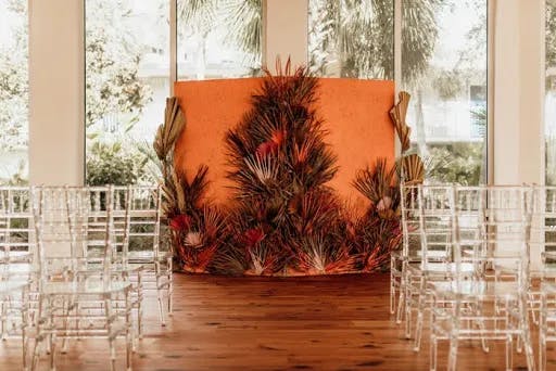 A beautifully decorated indoor wedding setup features clear acrylic chairs arranged in rows on a wooden floor, facing an orange backdrop adorned with vibrant tropical foliage and flowers. Large windows in the background reveal lush greenery outside.