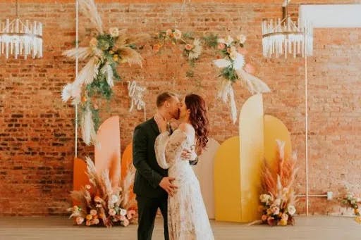 A couple shares a kiss at their wedding. They stand in front of a rustic brick wall adorned with vibrant floral arrangements and geometric decorations in warm tones. The bride wears a white dress, and the groom wears a dark suit.