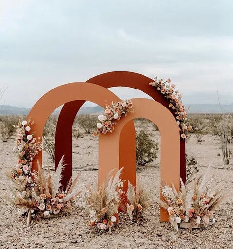A decorative wedding arch made of two large abstract arches is set in an arid desert landscape. The arches are adorned with floral arrangements featuring white, pink, and peach flowers, and pampas grass clusters are placed at the base of the structure.