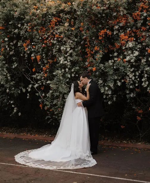 A bride and groom share a kiss in front of a wall covered in orange and white flowers. The bride wears a white dress with a long veil trailing behind her, and the groom is in a dark suit. They stand on a pavement with scattered petals.