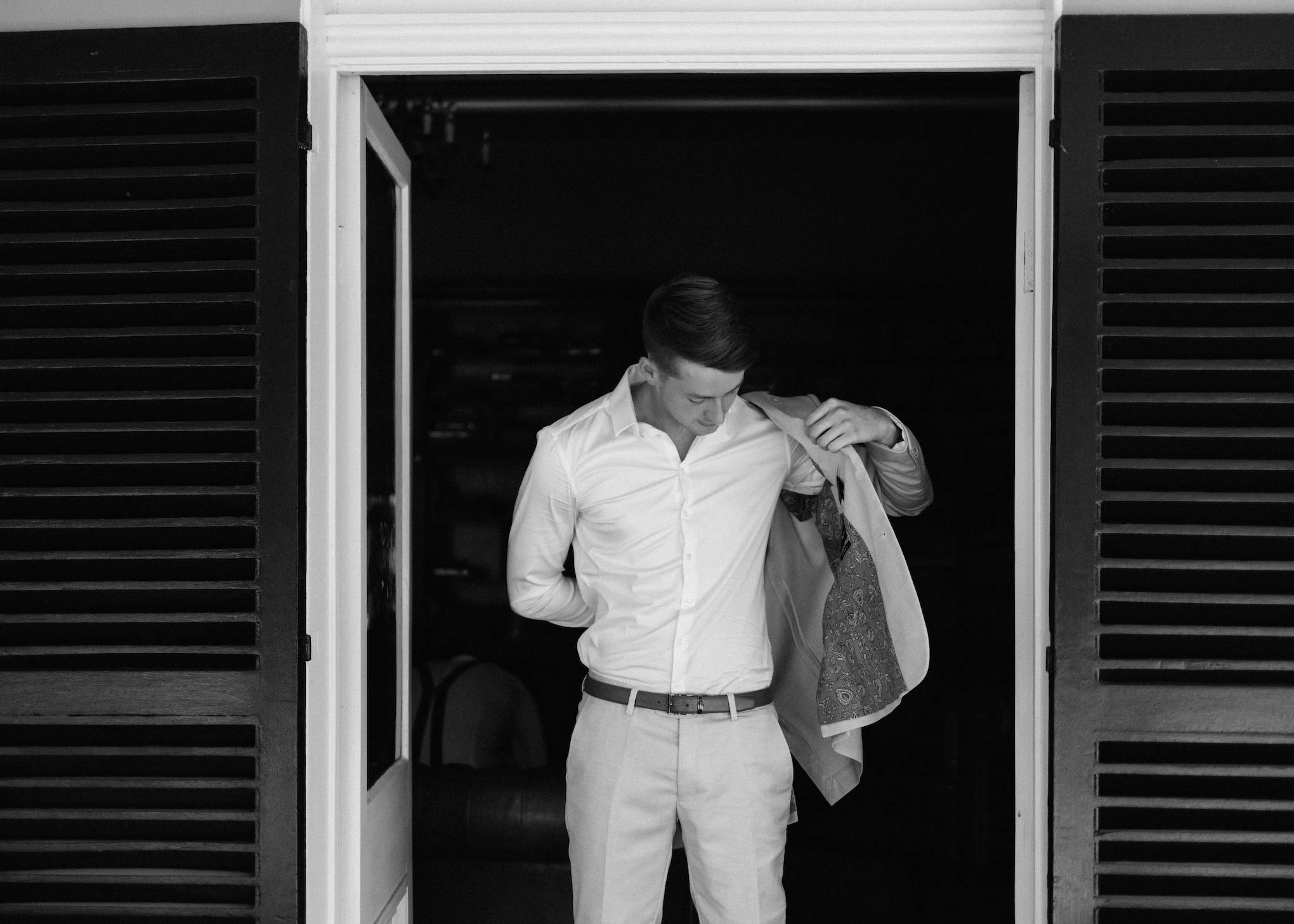 A man stands in an open doorway with dark shutters, putting on a blazer over his light-colored shirt and pants. He looks down and adjusts the jacket, preparing himself. The image is in black and white.