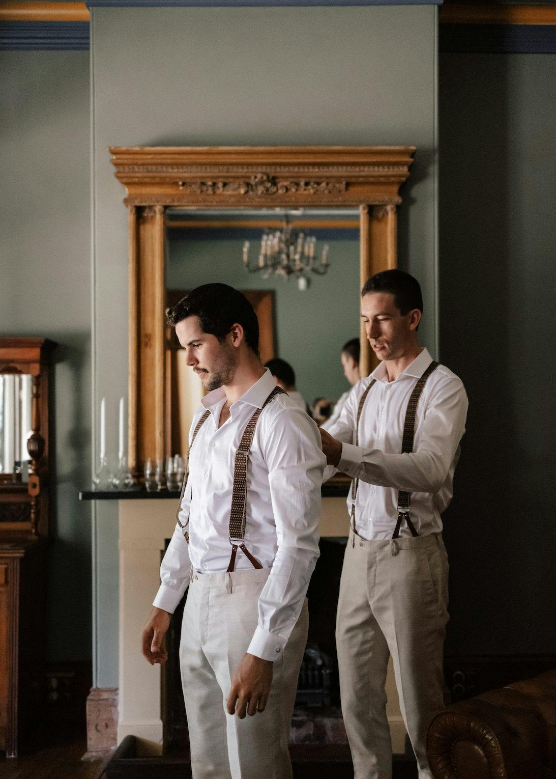 Two men in white shirts are getting ready in a well-decorated room with a large mirror and candles. One adjusts the other's suspenders, ensuring a neat appearance. The room has a classic and elegant decor with a fireplace and wooden furniture.