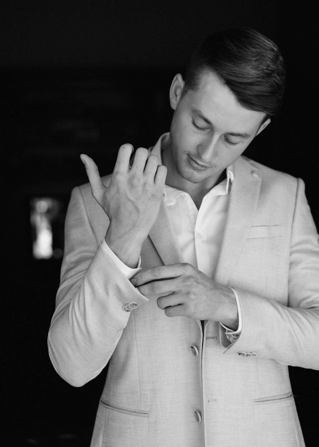 Black and white image of a man adjusting the button on the sleeve of his light-colored suit jacket. He is looking down with a focused expression in a dark background.