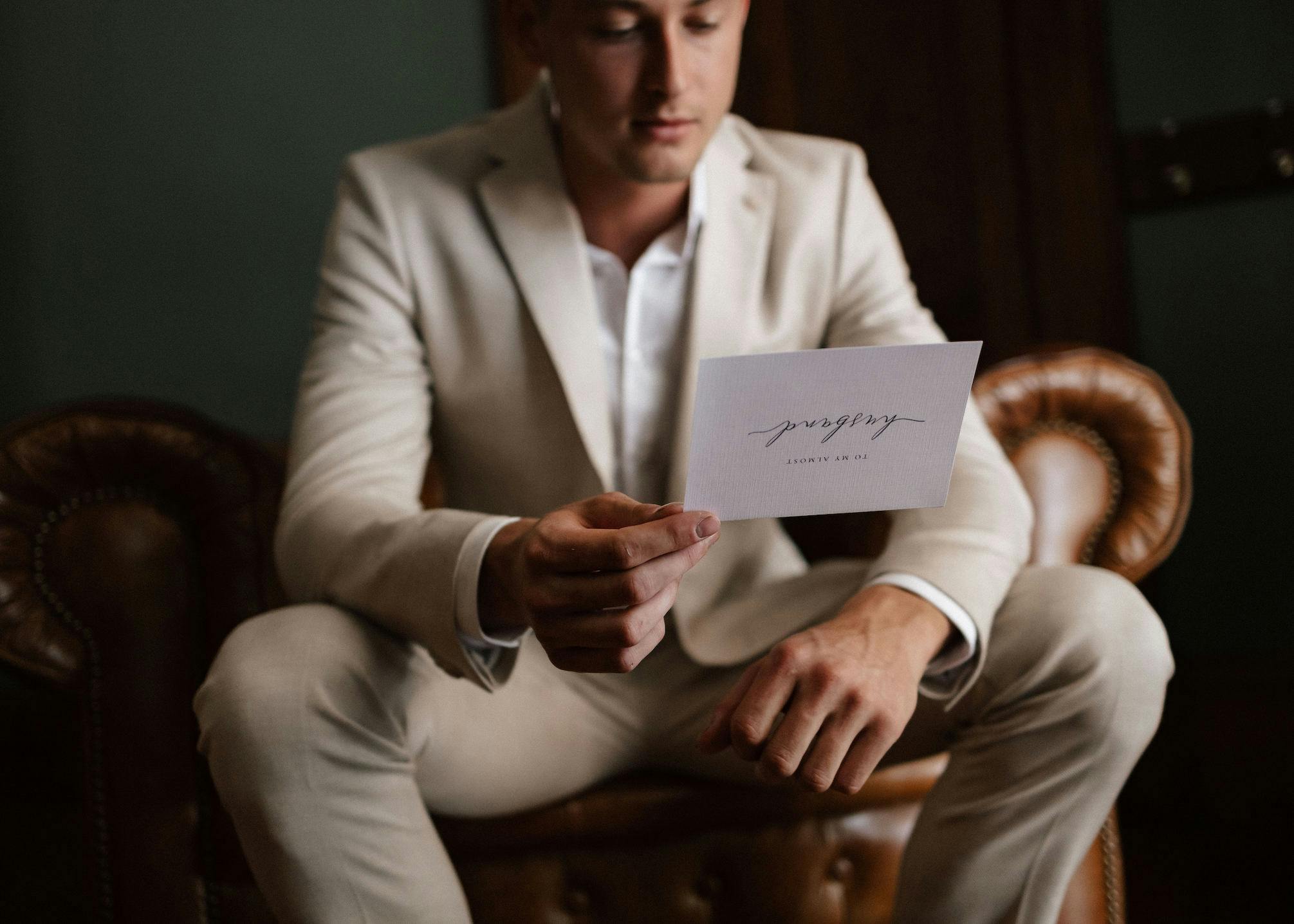 A person in a beige suit sits on a leather armchair, looking at a white card in their hand. The text on the card reads "journey" with a date below it. The background is dark and slightly out of focus.