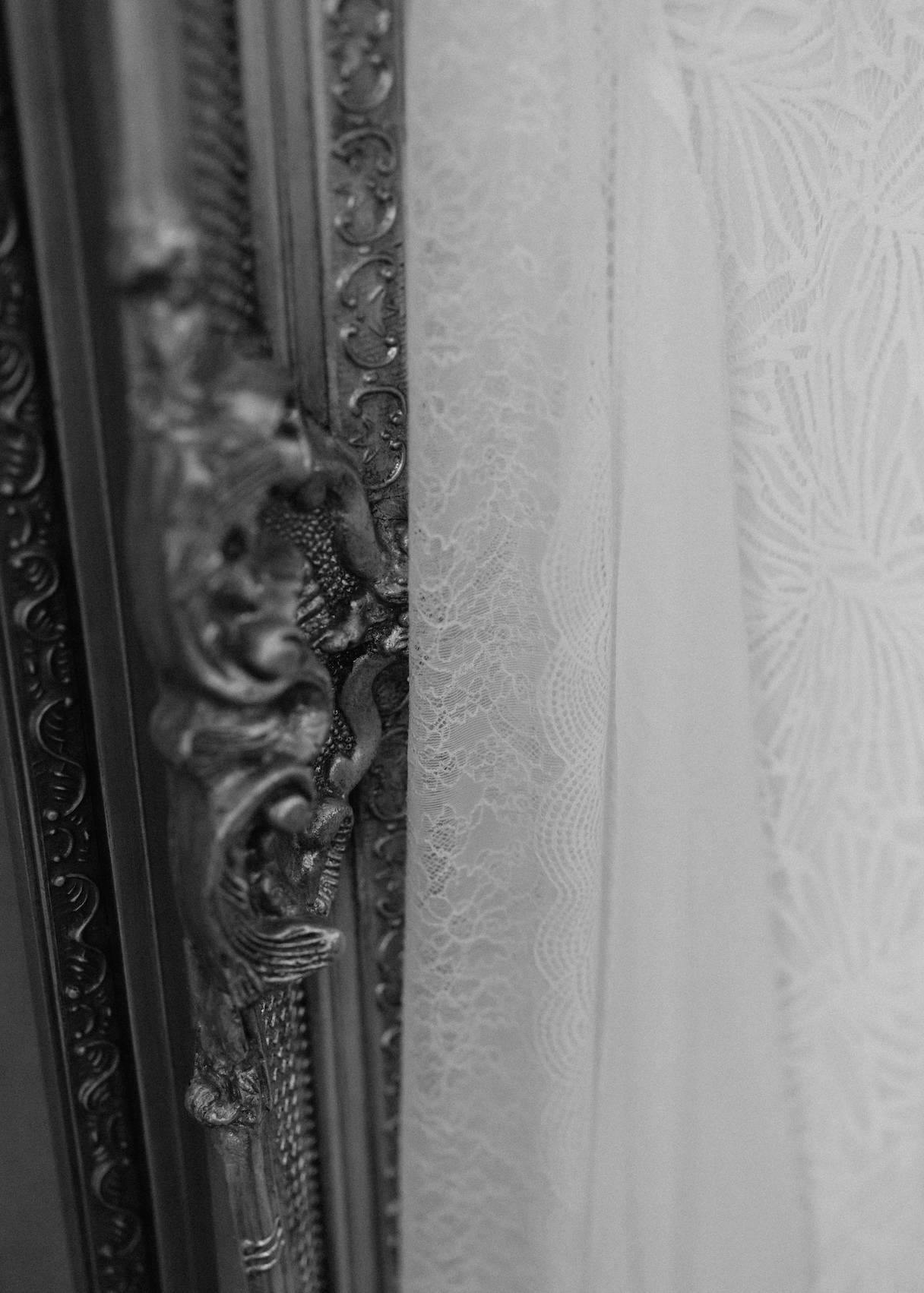Black and white close-up of an ornate vintage frame next to a delicate lace fabric. The frame features intricate carvings, and the lace shows a subtle floral pattern, emphasizing the textures and details of both materials.