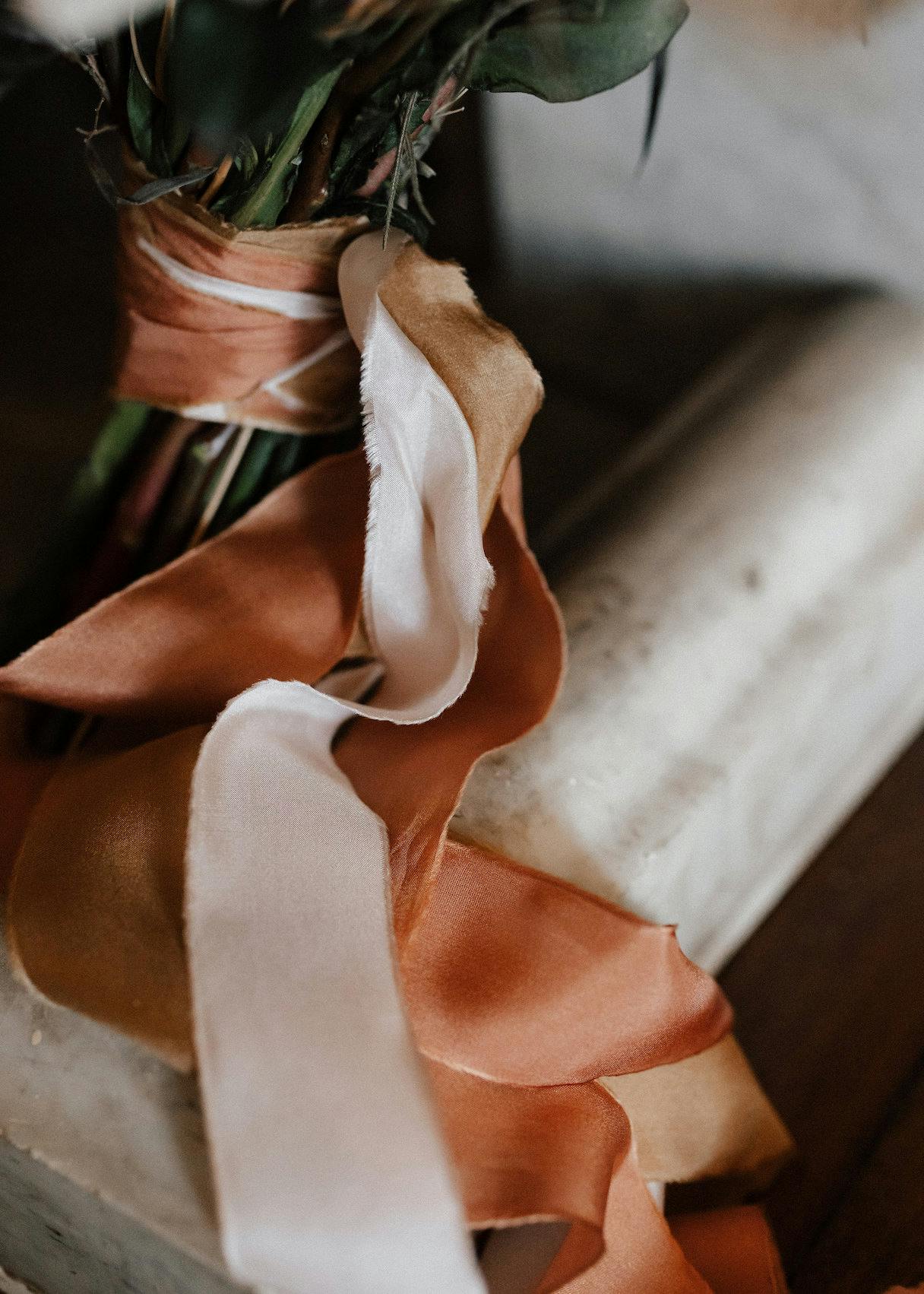 A close-up of a bouquet wrapped with orange, gold, and white ribbons. The ribbons are draped gracefully over a marble surface, creating a soft and elegant scene with natural textures and colors.