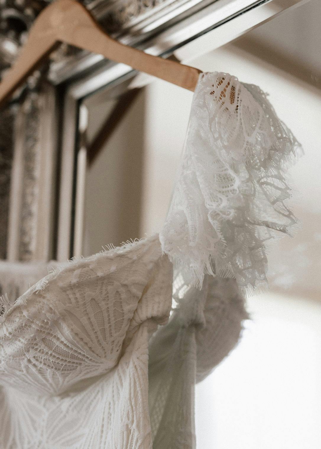 Close-up of a white lace wedding dress hanging on a wooden hanger in front of a mirror. The intricate lace detail and delicate fabric are emphasized, showcasing the craftsmanship of the garment. The soft lighting adds a romantic ambiance to the scene.