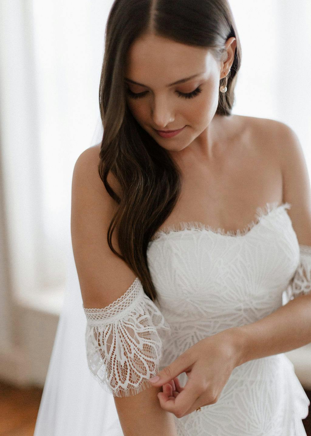 A bride with long brown hair adjusts the lace sleeve of her off-the-shoulder wedding dress. The dress features intricate lace detailing. She is looking down with a serene expression, standing indoors with a softly lit background.