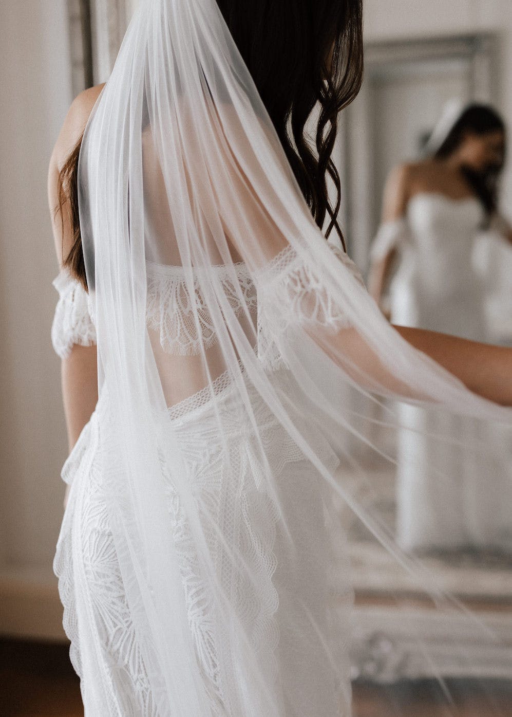 A bride wearing a white lace wedding dress and sheer veil stands with her back to the camera. Her reflection is visible in a large mirror. The room has soft lighting, giving the scene an ethereal feel.