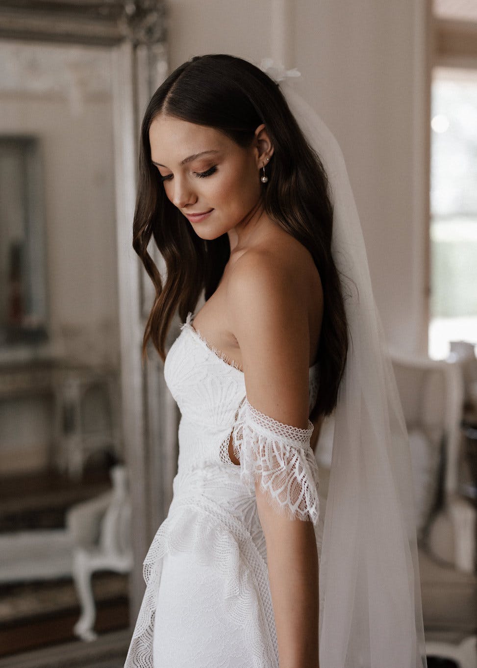 A bride with long dark hair stands in a softly lit room, wearing an elegant, off-shoulder lace wedding dress and a veil cascading down her back. She looks down with a serene expression, showing off her delicate pearl earrings.