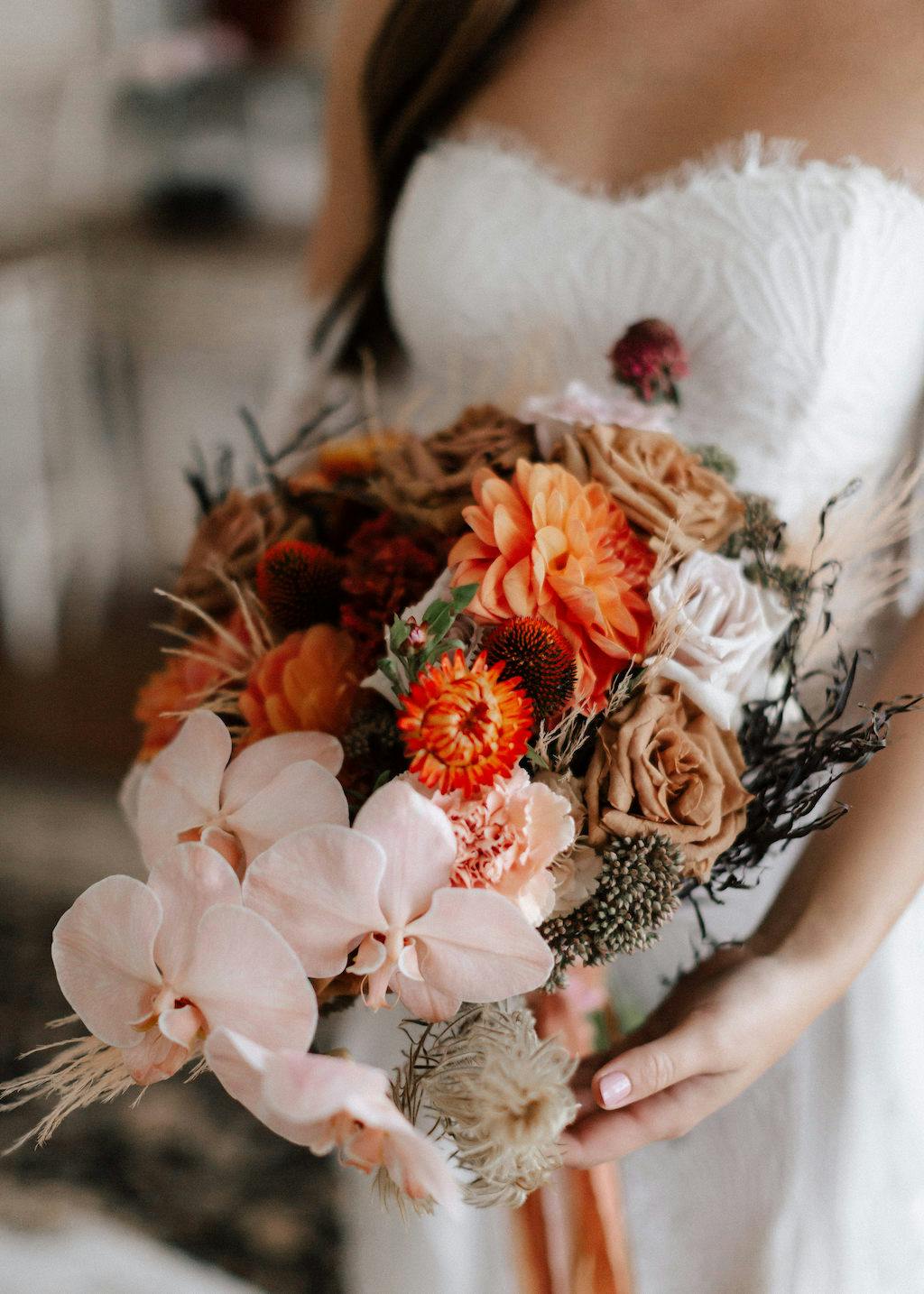 A bride wearing a strapless white dress holds a colorful bouquet featuring pink orchids, orange dahlias, and various other flowers with a mix of greenery and dried elements. The background is softly blurred, drawing focus to the bouquet.