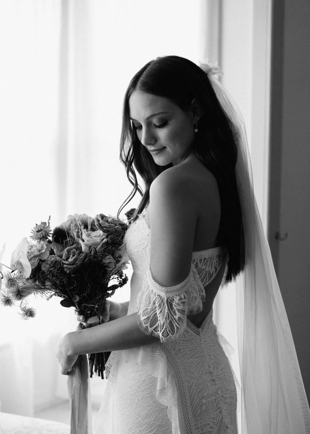 Black and white photo of a bride holding a bouquet of flowers. She is dressed in a lace wedding gown with off-the-shoulder sleeves and a long veil. She is looking down, showcasing the intricate details of her dress and bouquet in front of a softly lit window.
