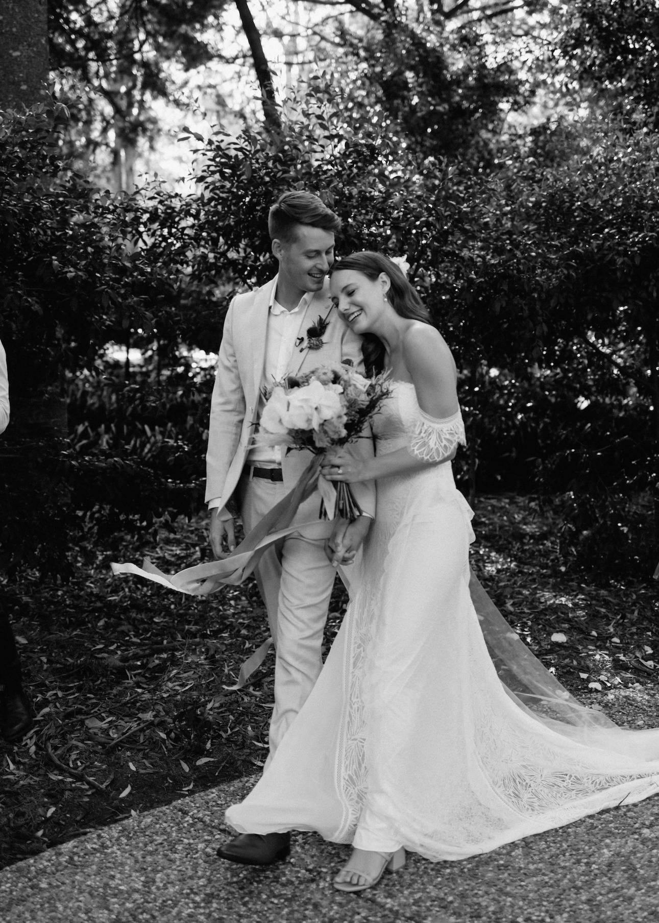 A couple is walking outside on a stone path surrounded by greenery. The groom, wearing a light-colored suit, is smiling at the bride, who is dressed in a long, elegant white gown, holding a bouquet of flowers and looking back at him. Both appear happy and joyful.