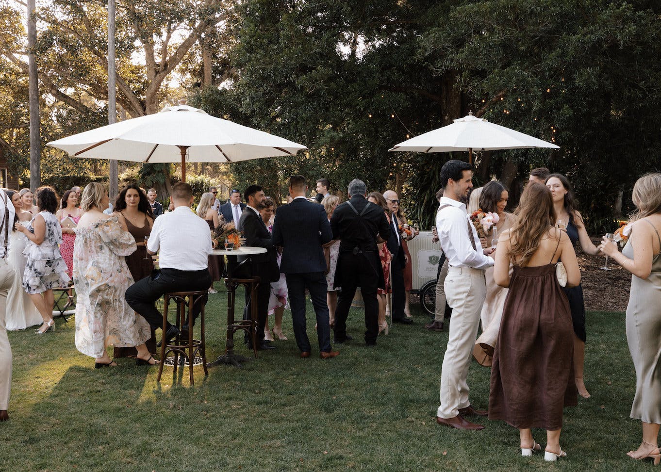 A group of people dressed in formal attire socializes at an outdoor event. They are gathered around white umbrellas and cocktail tables on a grassy area, with trees and string lights in the background. Some are holding drinks, while others are engaged in conversation.