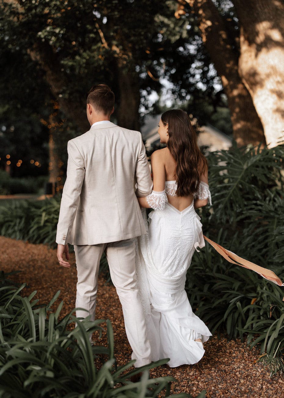 A bride and groom walk hand in hand down a garden path. The bride wears a white, off-the-shoulder wedding dress with lace details and a train, and the groom is dressed in a light beige suit. They are surrounded by lush greenery and trees, with lights glowing in the background.