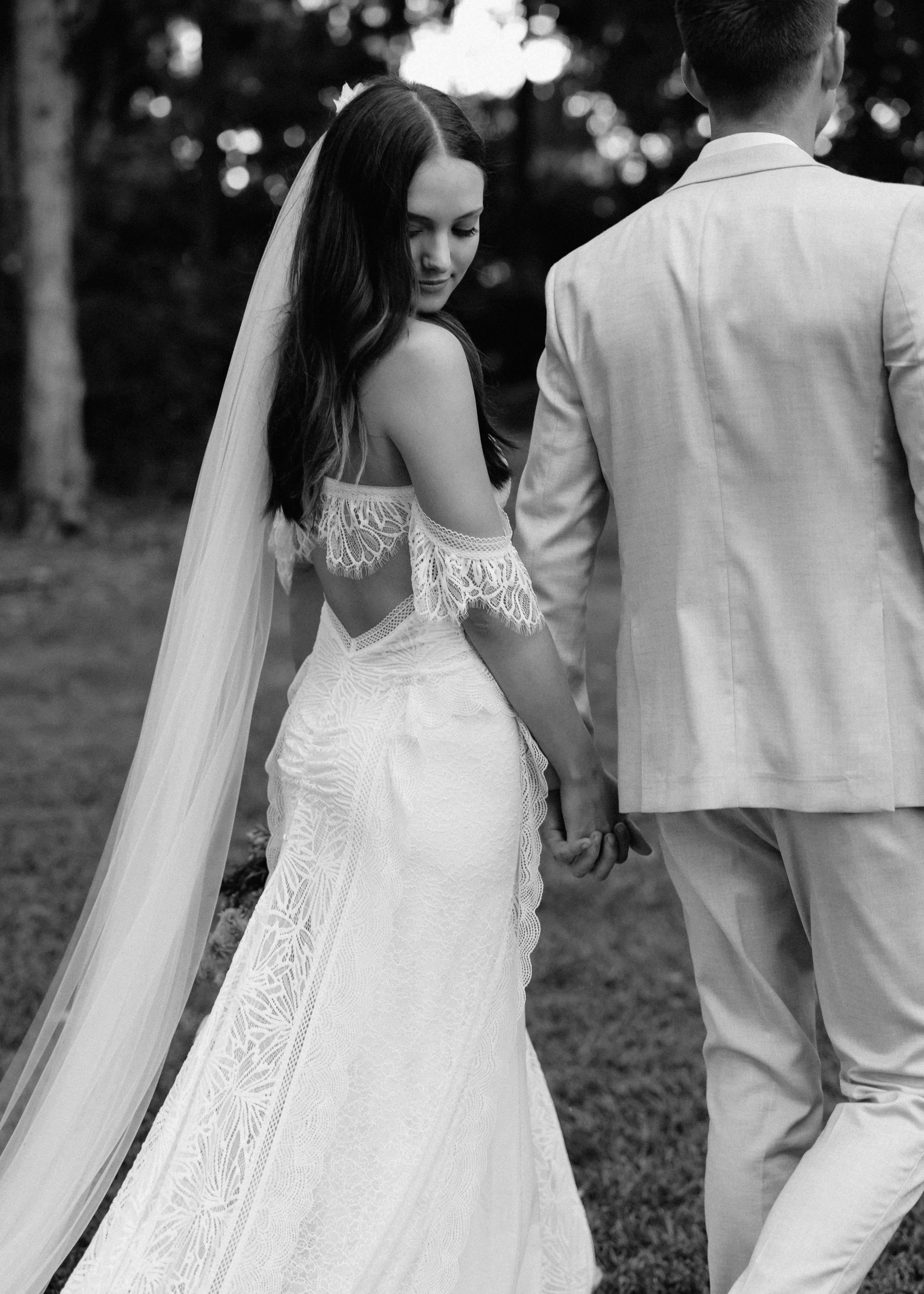 A bride and groom walk hand-in-hand through a grassy area. The bride, in a detailed lace gown and veil, glances back over her shoulder with a soft smile. The groom, dressed in a light-colored suit, faces forward. The monochrome image captures a serene, romantic moment.