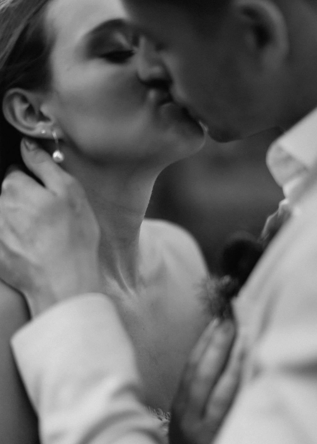 Black and white close-up photo of a couple sharing a tender kiss. The woman, wearing a strapless top and drop earrings, is gently holding the man's neck. The man, in a buttoned-up shirt, has his arm wrapped around her shoulder.