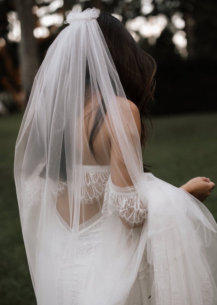 A bride with long, dark hair is photographed from behind, holding up her lace-embellished wedding gown. She wears a flowing white veil that drapes over her back and shoulders. The background consists of greenery, indicating an outdoor setting.
