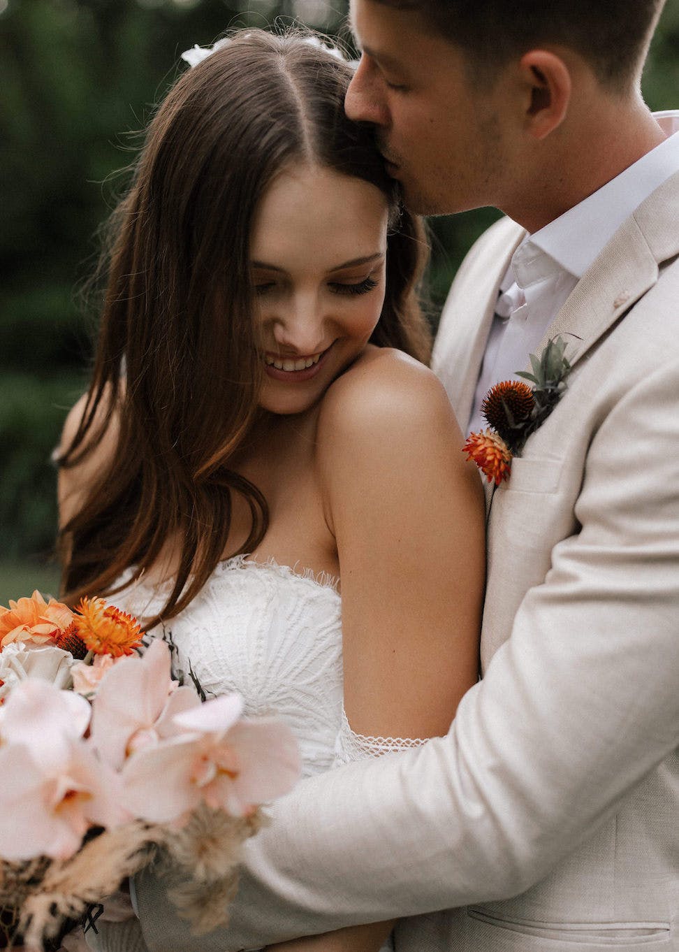 A groom in a light beige suit tenderly kisses the forehead of his smiling bride, who is wearing a strapless white dress and holding a bouquet of flowers with orange accents. They stand closely embraced, surrounded by greenery, radiating happiness and love.