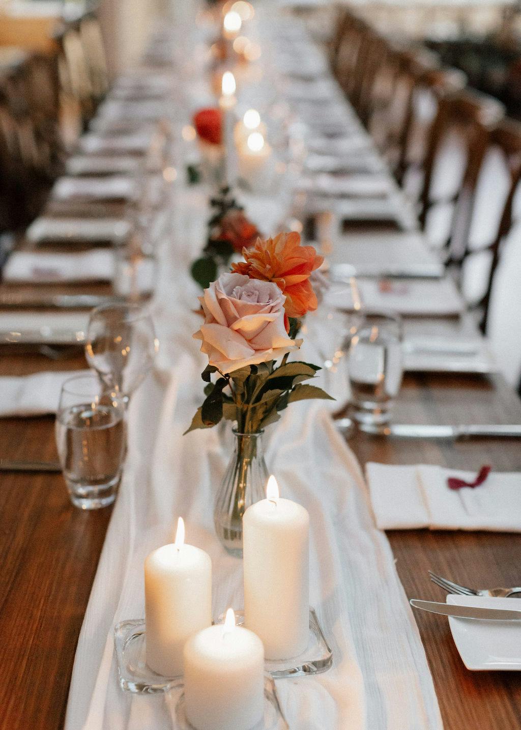 A long, elegantly set dinner table with wooden chairs. The table features a white runner, lit white pillar candles, and a centerpiece of fresh flowers in vases, including pink and orange blooms. Place settings include glassware, napkins, and flatware.