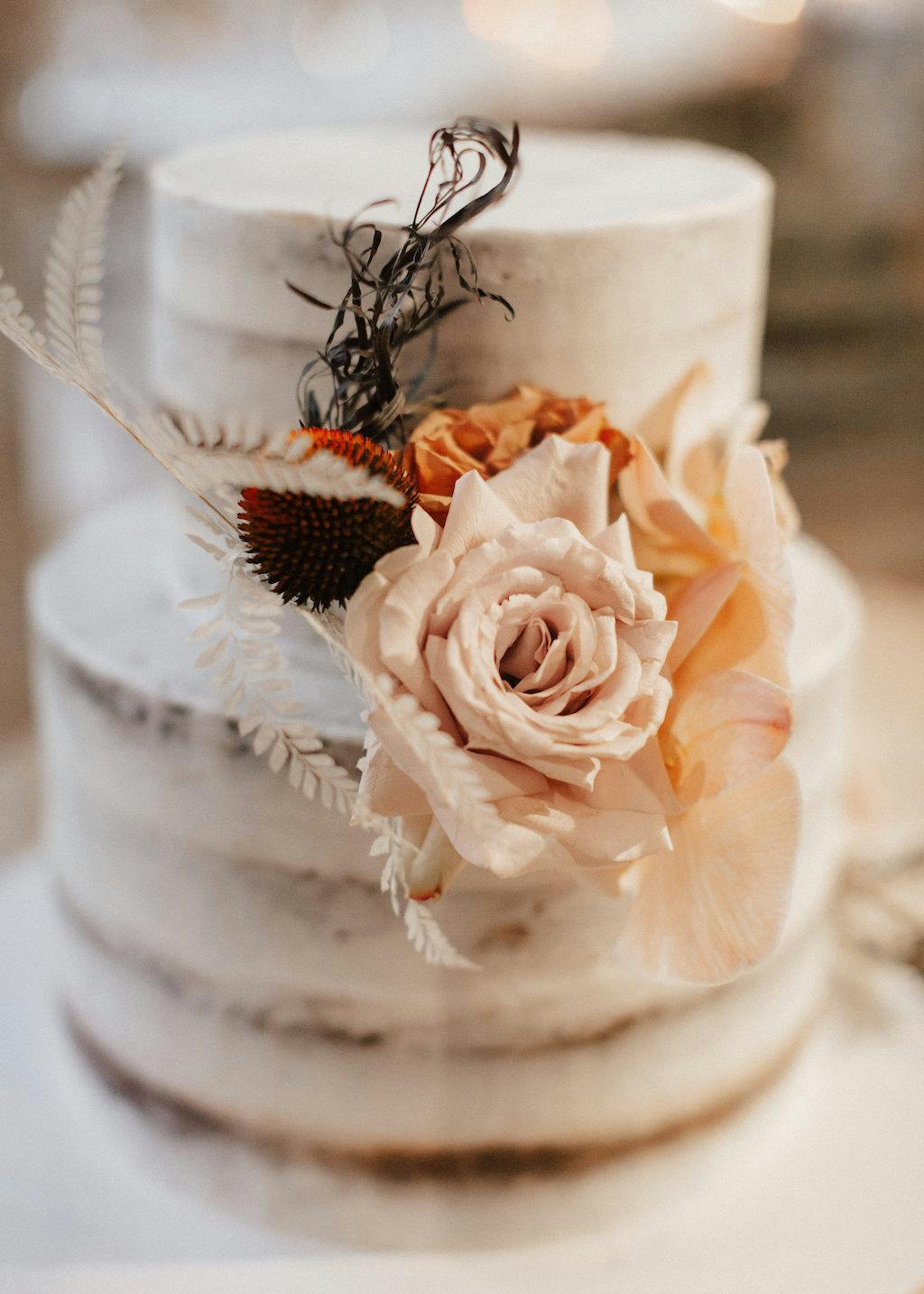 A two-tier, semi-naked wedding cake adorned with pale pink roses, dried orange flowers, and delicate white leaves, elegantly arranged in a natural, rustic style. The cake sits on a plain white surface with a softly blurred background.