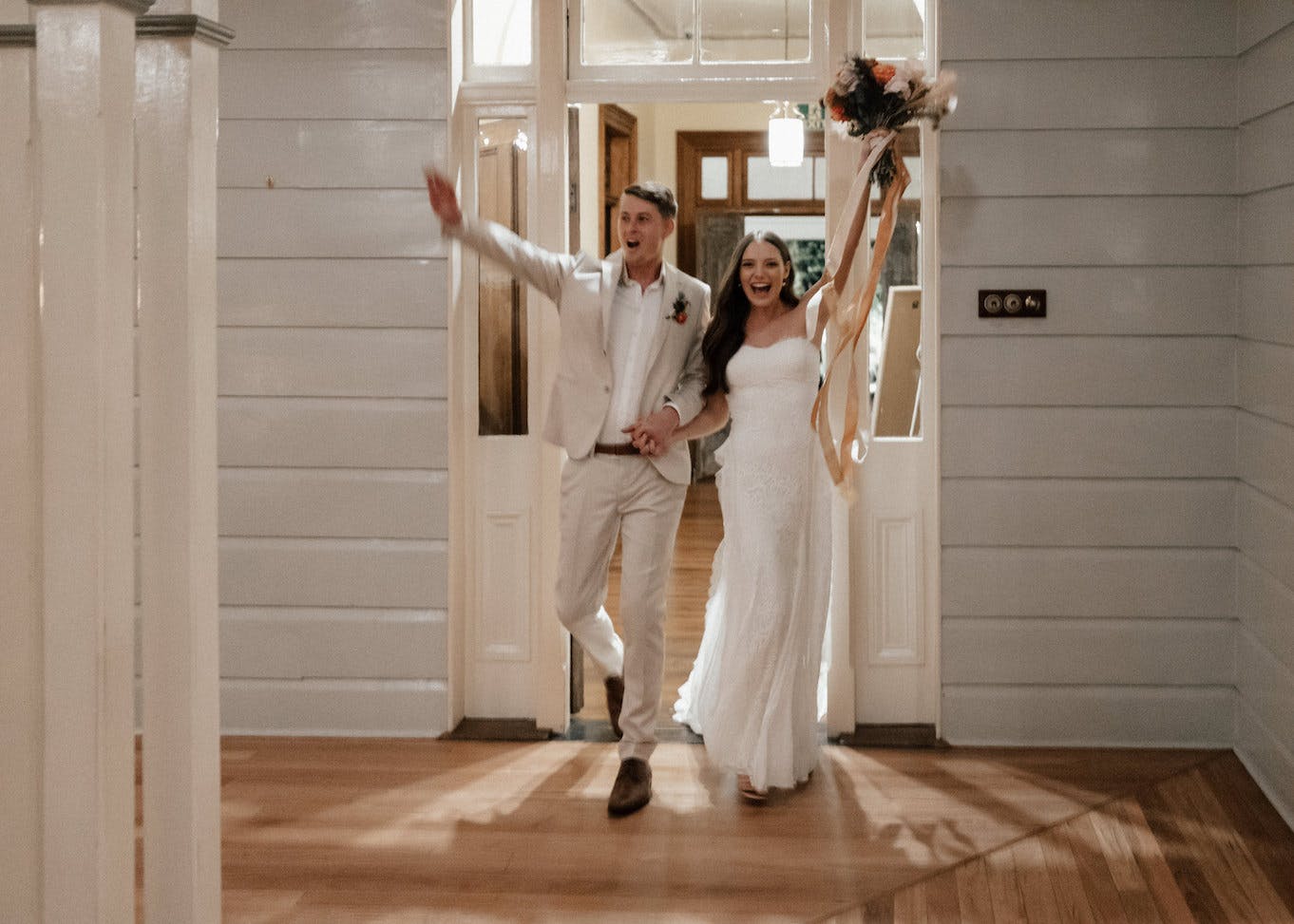 A joyful bride and groom are walking hand in hand through a doorway. The groom, in a light-colored suit, waves happily, while the bride, in a white gown, holds a bouquet up high with ribbons flowing. They are smiling and appear to be celebrating their wedding.