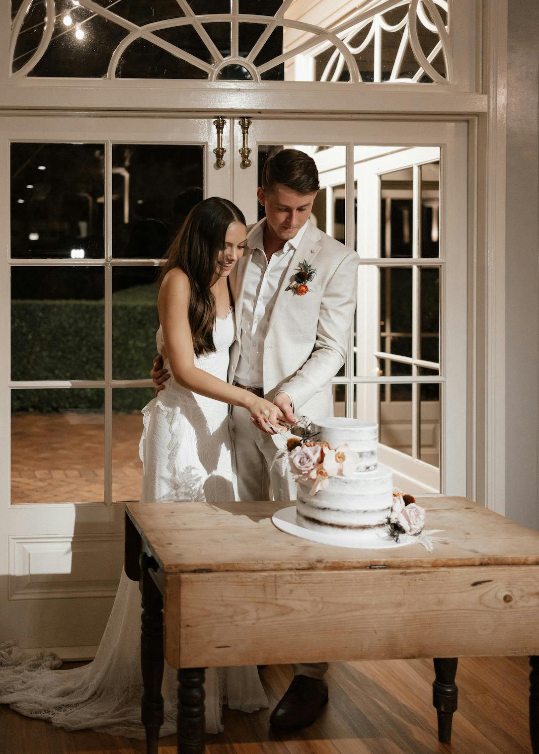 A couple in wedding attire stands together, cutting a tiered white cake decorated with pink flowers. They are in front of a large, ornate window, and the setting appears elegant and intimate. The bride wears a white dress, and the groom is in a light-colored suit.