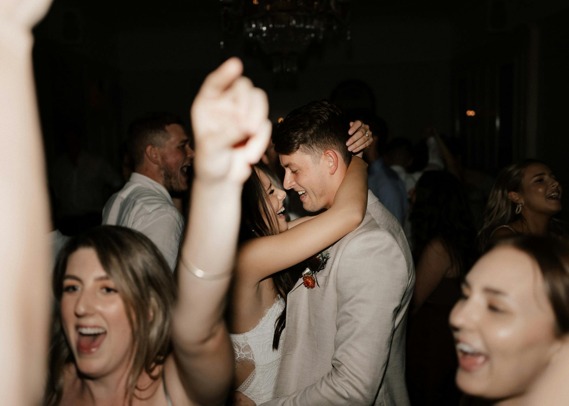 A couple is joyfully dancing and embracing on a crowded dance floor. People around them are cheering and dancing, visibly enjoying the moment. The atmosphere is lively and celebratory. The background is dimly lit, with a chandelier visible above.