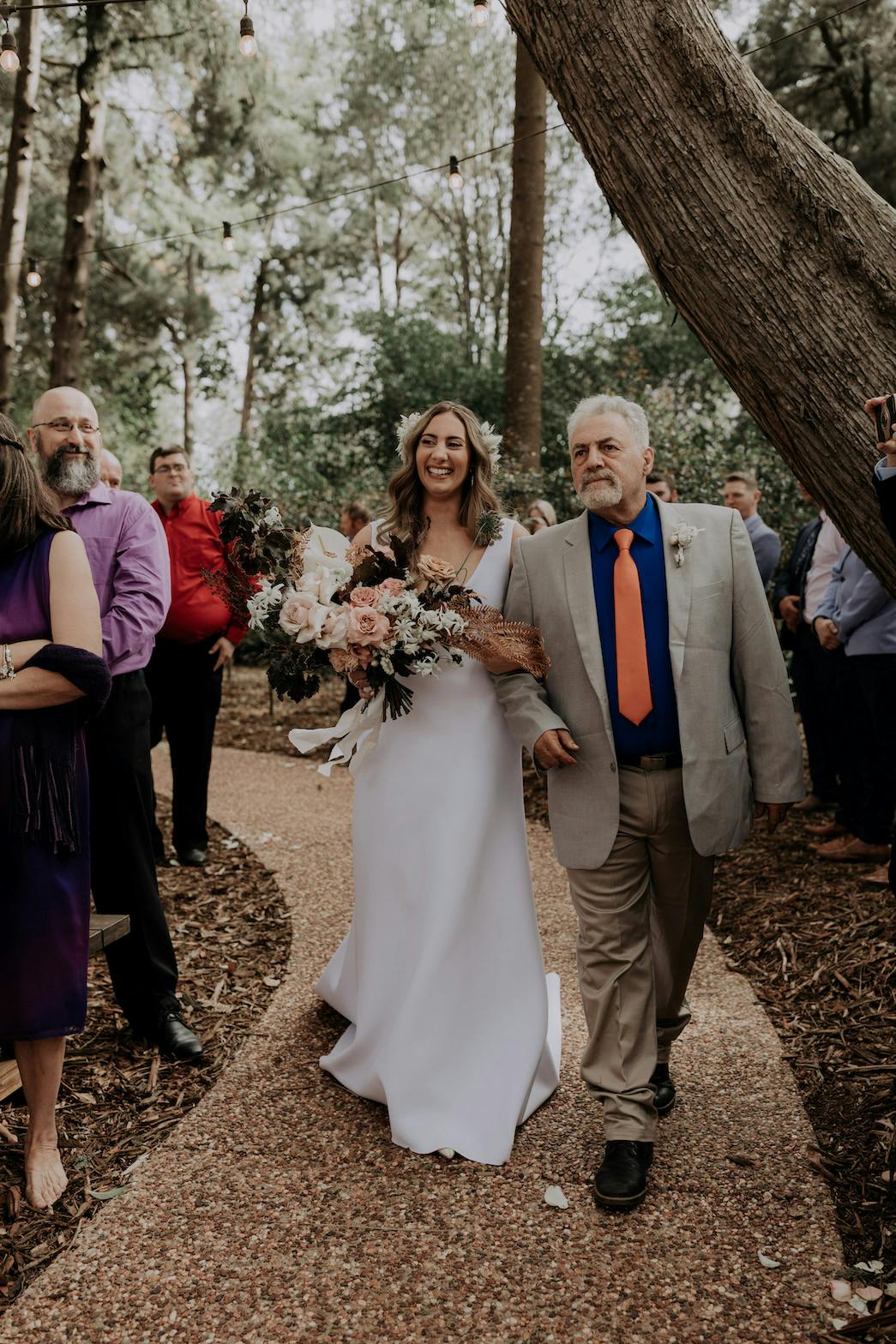 A bride in a white dress and veil walks down a wooded outdoor aisle, holding a large flower bouquet and accompanied by an older man in a grey suit with a blue shirt and orange tie. Guests in colorful attire stand on either side observing the scene.