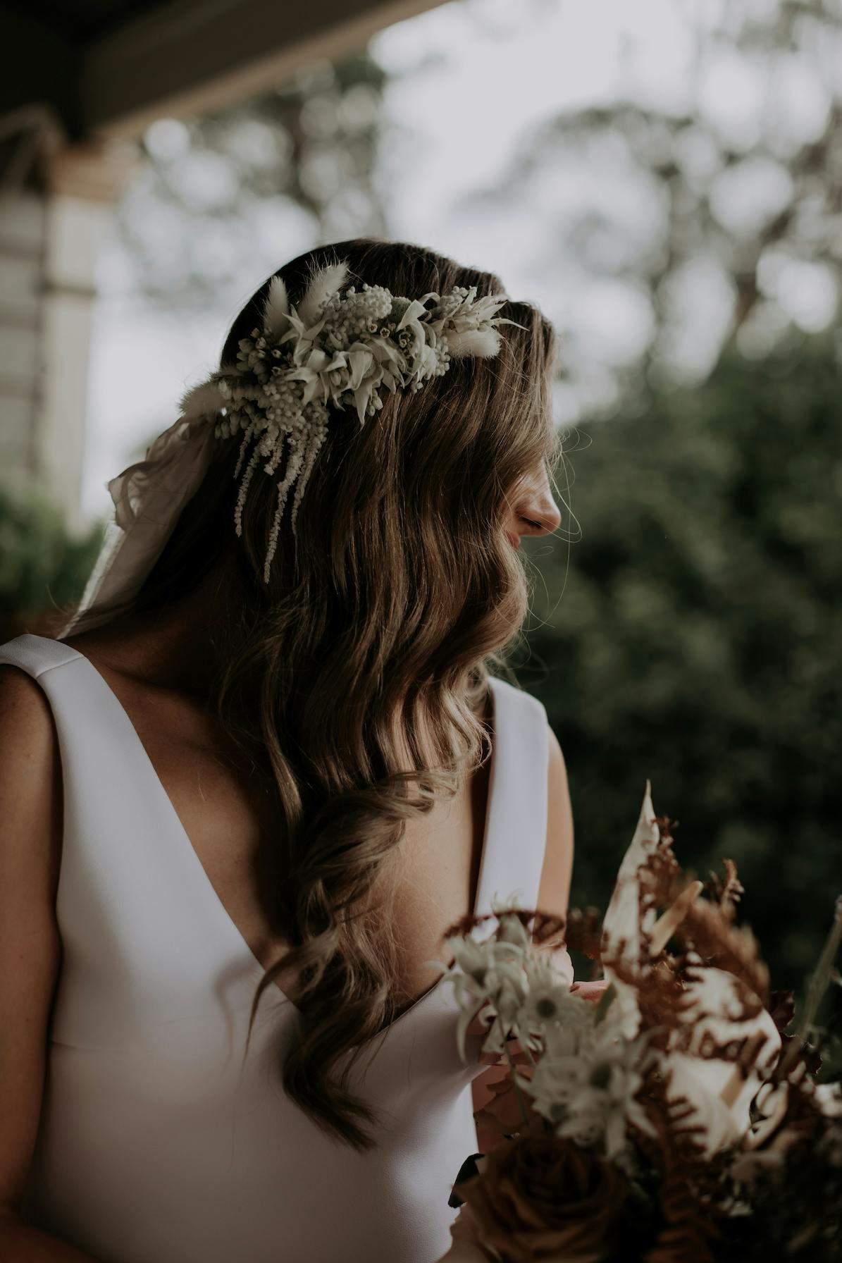 A bride with long, wavy brown hair adorned with a floral headpiece looks to the side. She is dressed in a simple white gown and holds a bouquet featuring white and brown flowers. The background is a blurred outdoor scene with greenery.