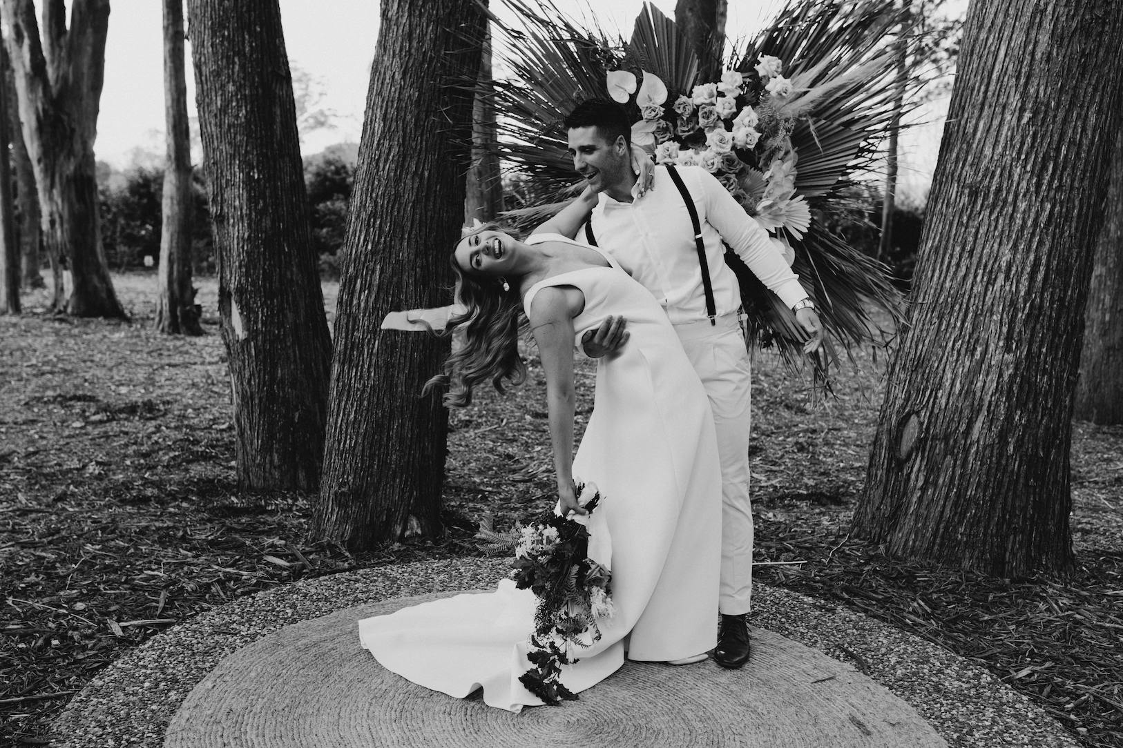 A black-and-white photo of a joyful bride and groom dancing. The groom, wearing suspenders, dips the bride, who is in a long, elegant dress and holding a bouquet of flowers. They are surrounded by tall trees and a decorative floral arrangement.