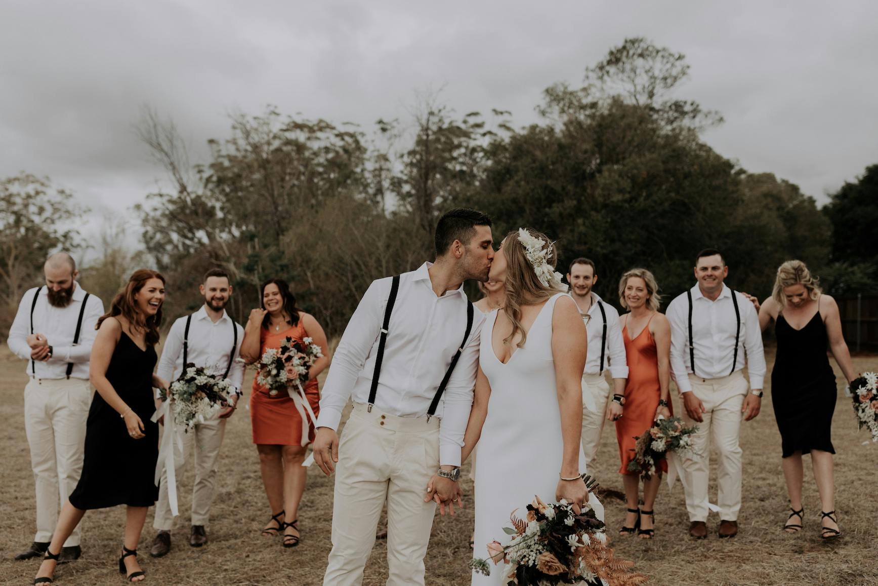 A bride and groom kiss while holding hands, surrounded by their joyful wedding party outdoors. The groom wears a white shirt with suspenders and light-colored pants; the bride wears a white dress. The bridesmaids wear black and orange dresses, and the groomsmen are in white shirts with suspenders.