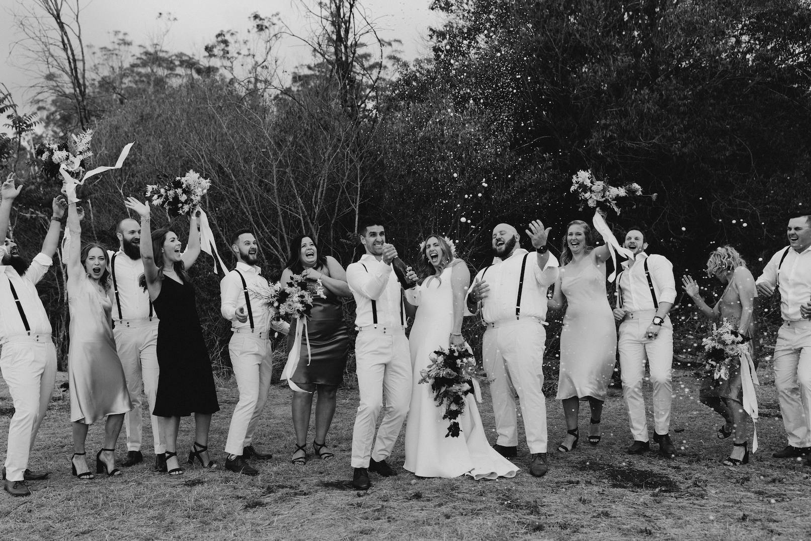 A black-and-white photo captures a joyful bridal party celebration. The bride and groom stand in the center, surrounded by bridesmaids and groomsmen, all cheering and holding bouquets. Confetti fills the air as everyone smiles and laughs. Trees form the background.