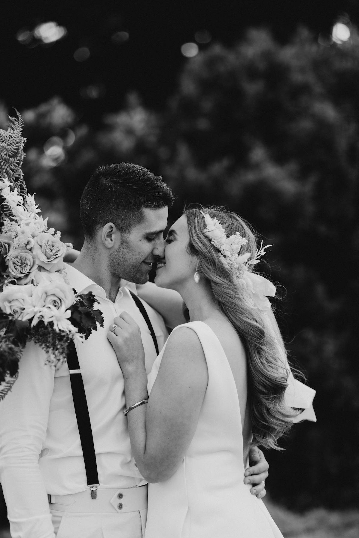 A black-and-white photo of a newlywed couple standing close, forehead to forehead, both smiling. The groom wears suspenders and a white shirt, holding a bouquet over his shoulder. The bride, adorned with a floral headpiece, has her arms around his neck.