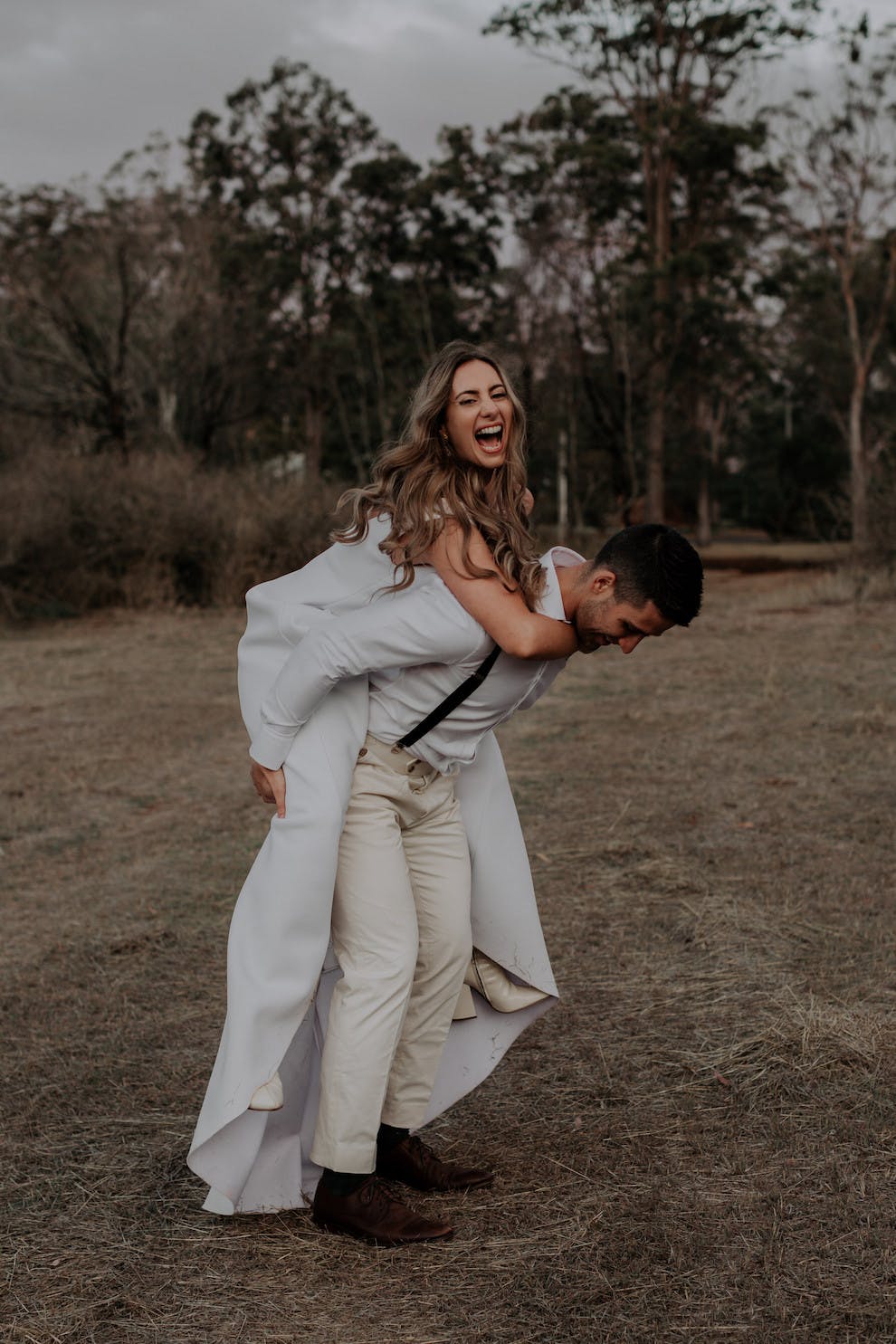 A joyful couple is outdoors, with the woman playfully piggybacking on the man. She is laughing, and they both look happy and carefree. The man is wearing suspenders and white pants, while the woman is in a white dress. Trees and grassy ground are in the background.