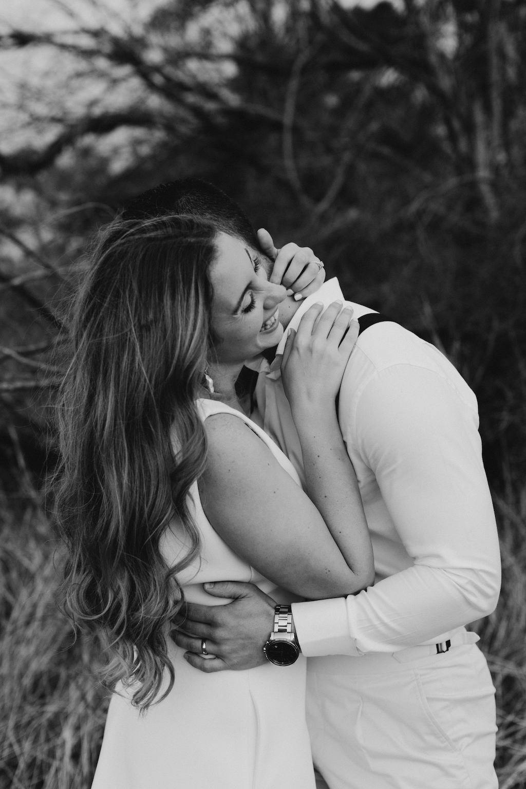 A couple shares a close, affectionate moment outdoors. The woman, with long hair and wearing a sleeveless dress, touches the face of the man, who is dressed in a white shirt and suspenders. They appear happy and intimate, standing amidst nature. Black and white photo.