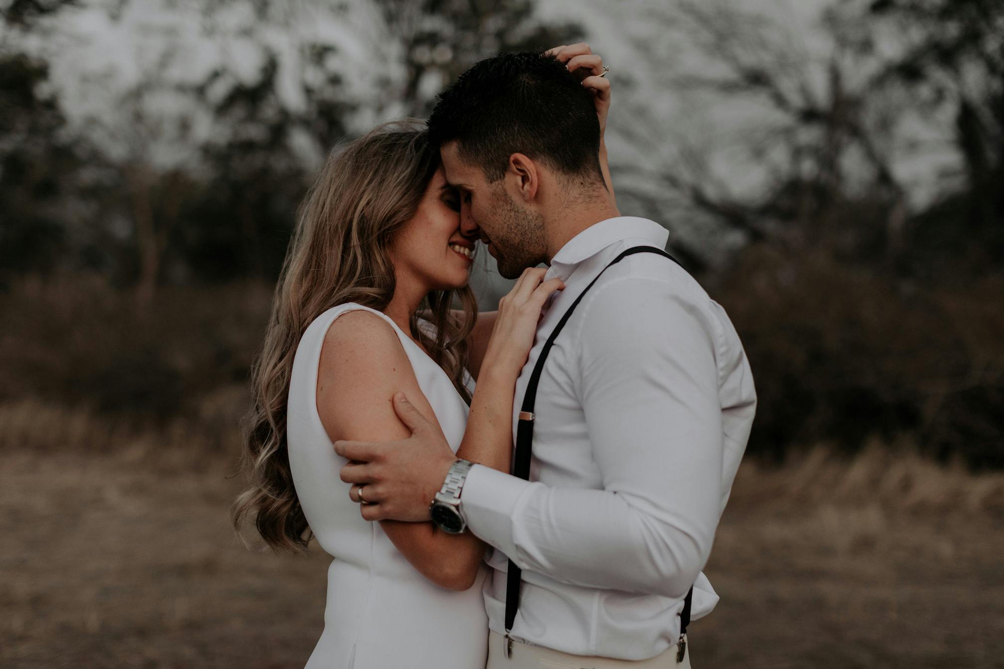 A couple dressed in white embraces closely in an outdoor setting with barren trees in the background. The woman has long, wavy hair, and the man wears suspenders. They share an intimate moment with closed eyes, their faces gently touching.