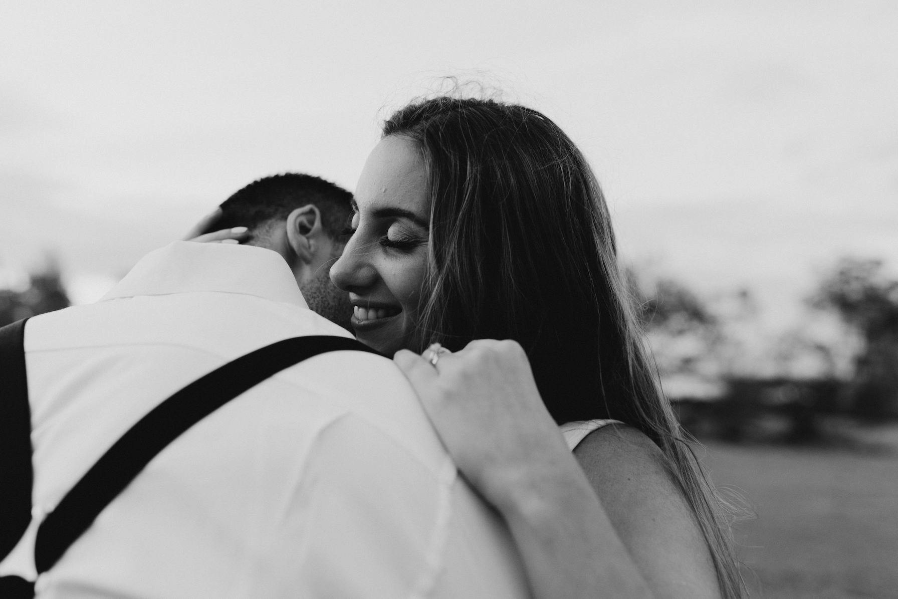 A black-and-white photo shows a couple embracing outdoors. The woman, smiling with her eyes closed, rests her head on the man's shoulder. The man, facing away from the camera, wears a white shirt with suspenders. The background is blurred trees and open sky.