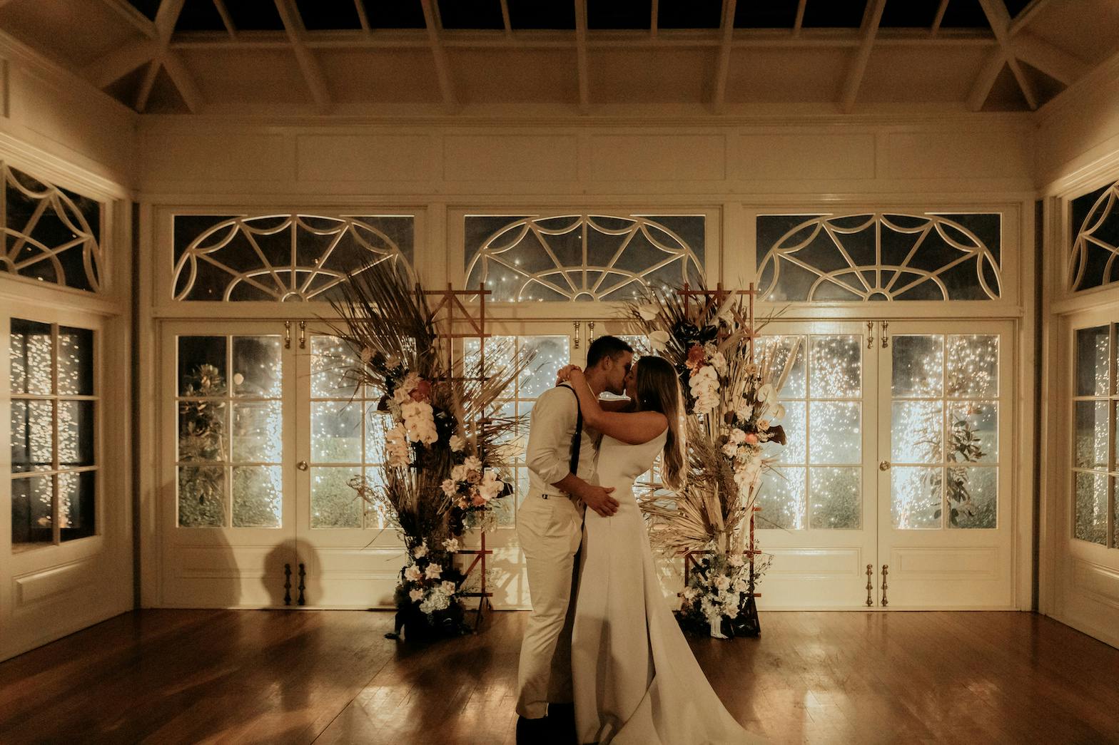 A newly married couple shares a kiss in an elegant, softly lit room. They are surrounded by floral arrangements and delicate lights, standing in front of large windows with intricate designs. The bride wears a long white dress, and the groom is in light attire.