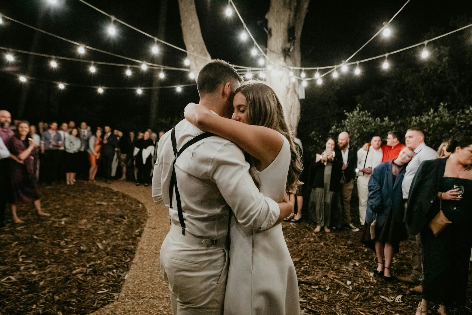 A couple shares a romantic dance under string lights at night during an outdoor event. The man is dressed in light-colored clothes with suspenders, and the woman is wearing a white dress. A crowd of onlookers stands in the background, watching and celebrating.