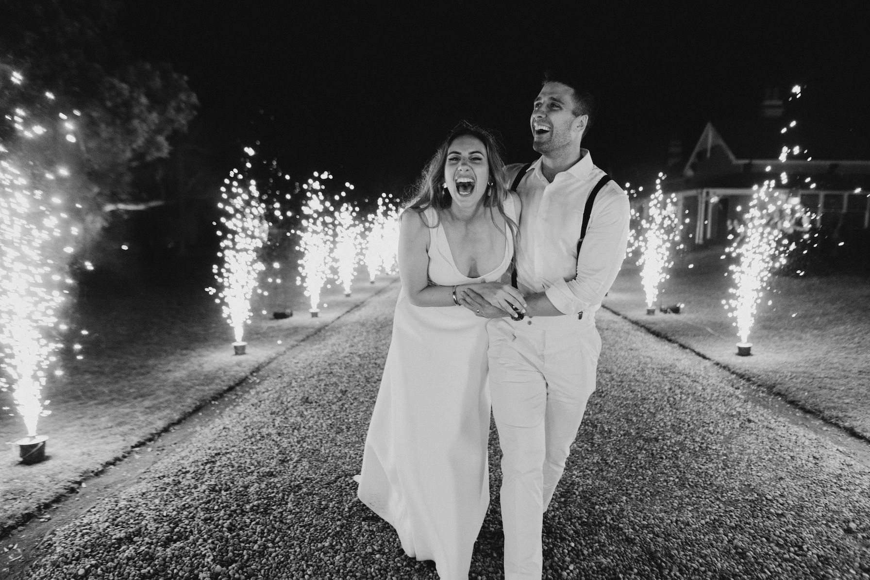 A couple dressed in formal attire joyfully laughs while holding hands and walking through a path lined with sparkling fireworks at night. The woman wears a gown and the man is in a shirt and suspenders. The background is dark, enhancing the bright fireworks' display.