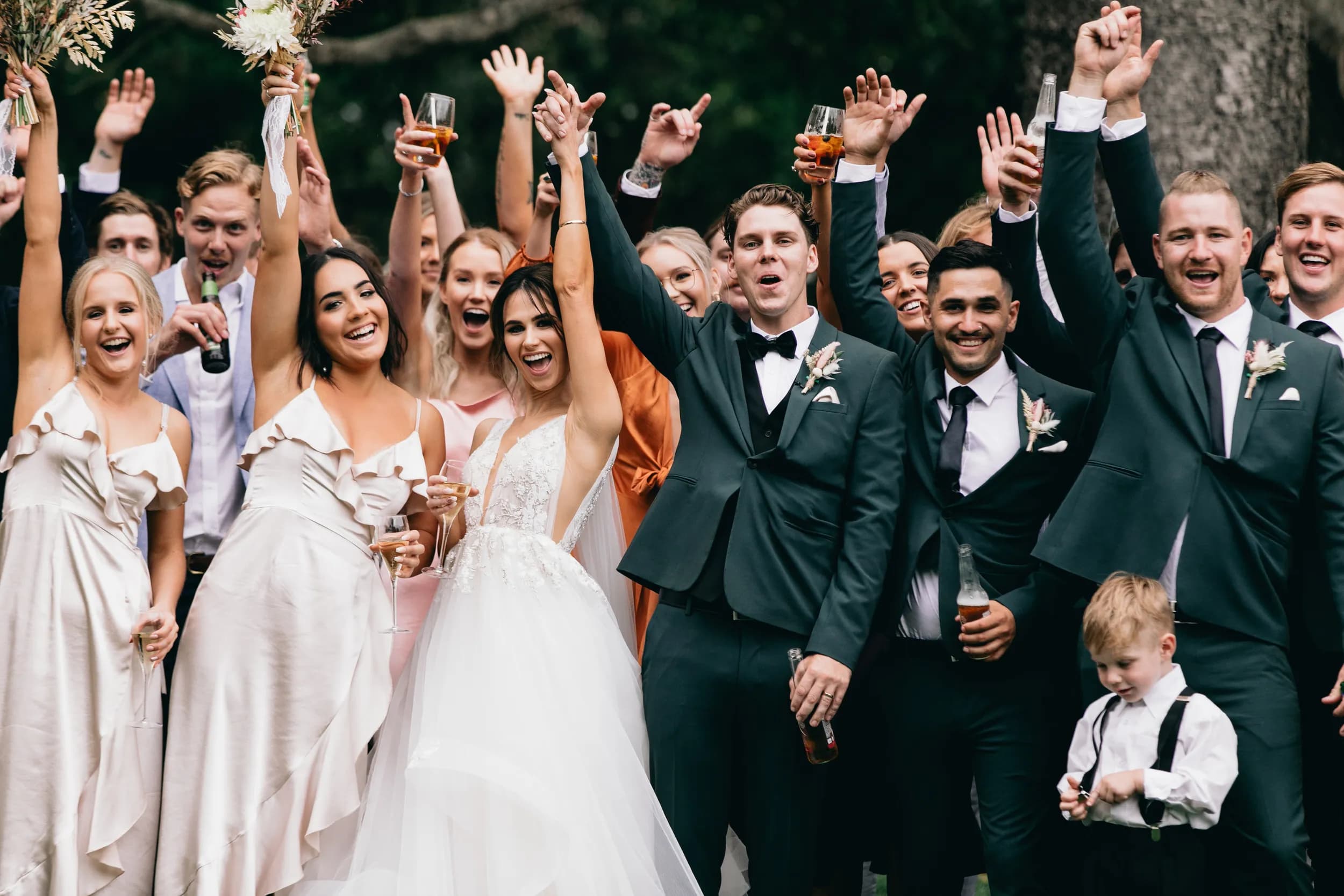 A large group of people, including a bride and groom, celebrate at an outdoor wedding. Everyone is dressed formally, smiling, and raising their arms in excitement. The bride and bridesmaids wear white dresses, while the groom and groomsmen wear dark suits.