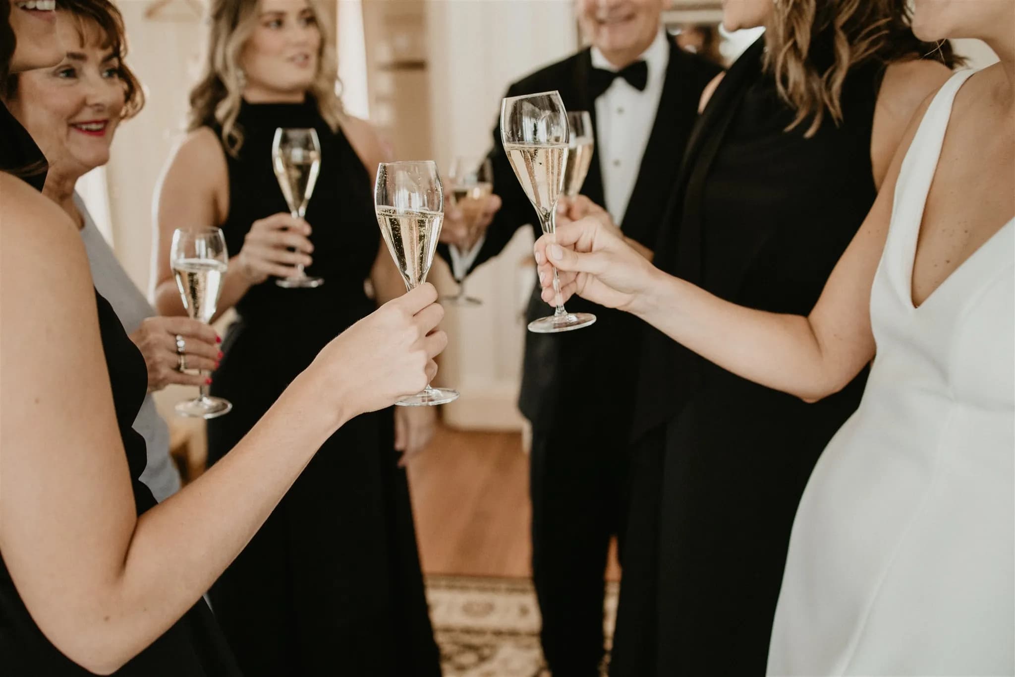 A group of people dressed in elegant attire, including women in black dresses and a man in a tuxedo, raise champagne glasses in a celebratory toast. The atmosphere is festive, with warm lighting and a cozy interior setting.