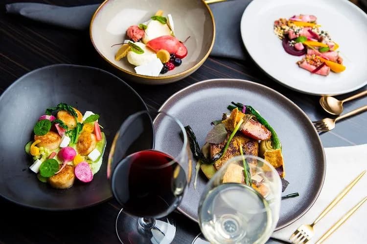 A table set with four elegant plates of gourmet food, featuring a variety of colorful vegetables and meats, and two glasses of wine—one red and one white. The table setting includes gold-colored cutlery and a dark, modern tablecloth.